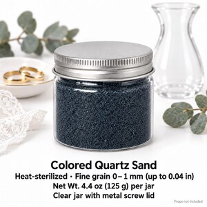 Closed clear jar of dark navy blue colored quartz sand with a silver metal screw lid, next to two gold wedding rings on a white dish, a clear glass vase, eucalyptus and white lace. Text: Colored Quartz Sand, heat-sterilized, fine grain 0-1 mm (up to 0.04 in), Net Wt. 4.4 oz (125 g) per jar, clear jar with metal screw lid, props not included.