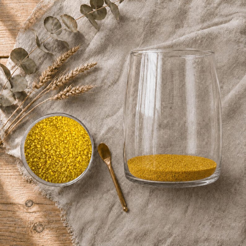 Small dish of bright yellow craft sand, a gold spoon, and a glass vase with a thin yellow sand layer, beside wheat and eucalyptus