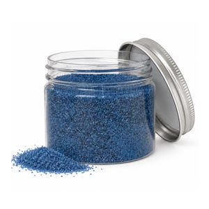 Open clear jar of medium blue colored quartz sand with its silver metal screw lid leaning behind it and a small pile of the same sand spilled in front, showing the fine grain texture, on a white background.