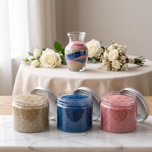 Three open clear jars of colored quartz sand in natural beige, royal blue and dusty pink, with silver lids leaning behind them, on a marble counter. In the background, a glass vase with wavy beige, blue and pink sand layers stands on a round table with white roses and a small bouquet.