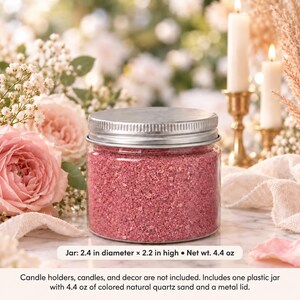 Closed clear jar of rose pink colored quartz sand with a silver metal screw lid on a marble table, surrounded by pink roses, baby's breath, lit white candles in brass holders and cream fabric. Text: Jar: 2.4 in diameter x 2.2 in high, Net wt. 4.4 oz. Candle holders, candles, and decor are not included. Includes one plastic jar with 4.4 oz of colored natural quartz sand and a metal lid.