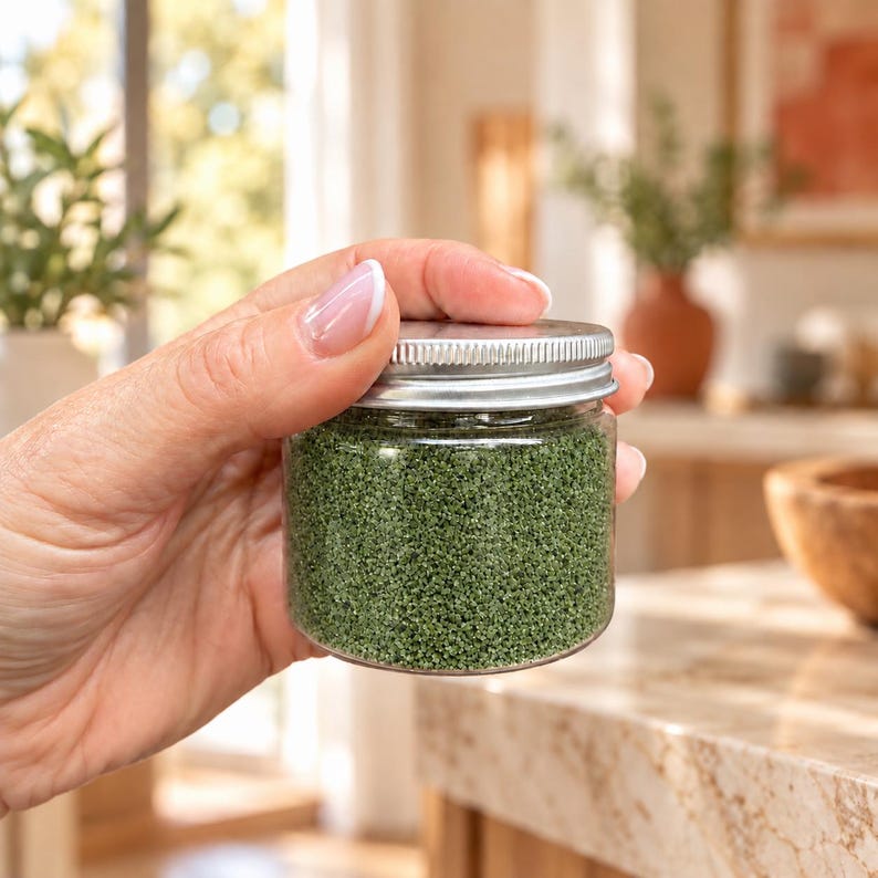 Hand holding a small closed jar of Forest Moss green craft sand with a silver lid to show size, in a bright kitchen