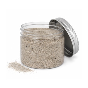Open clear jar of natural light beige quartz sand with its silver metal screw lid leaning behind it and a small pile of the same sand spilled in front, showing the fine grain texture, on a white background.