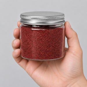 Hand holding a closed clear jar of Wild Berry red quartz sand with a silver metal screw lid, showing the jar's small size, on a light grey background.