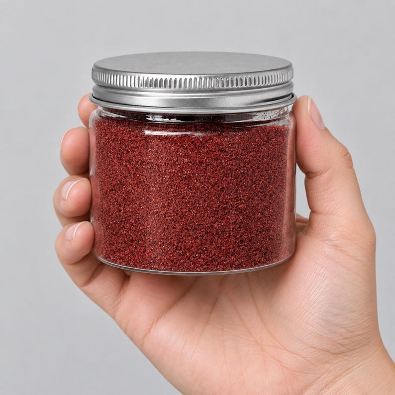 Hand holding a closed clear jar of Wild Berry red quartz sand with a silver metal screw lid, showing the jar's small size, on a light grey background.