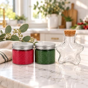 Op de afbeelding: Twee cilindrische glazen potten met zilverkleurige metalen deksels, één gevuld met rood korrelig materiaal en één met groen korrelig materiaal, naast een doorzichtige glazen fles in stervorm met een cilindrische kurk.