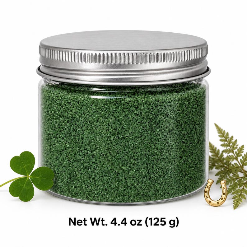 Closed clear jar of Forest Moss colored quartz sand in a deep forest green, with a silver metal screw lid, next to a three-leaf clover, a small gold horseshoe and a fern sprig, with text: Net Wt. 4.4 oz (125 g).
