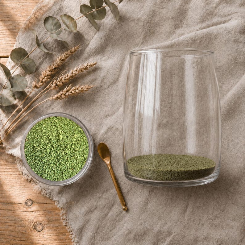 Small dish of green craft sand, a gold spoon, and a glass vase with a thin green sand layer, beside wheat and eucalyptus