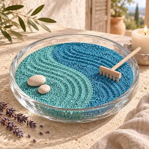 Glass dish zen garden with raked swirls of aqua and blue sand, two smooth stones, and a small wooden rake, next to a candle and lavender