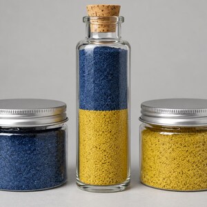 Tall cork-stoppered glass bottle filled with two layers of quartz sand in the colors of the Ukrainian flag, Golden Saffron yellow at the bottom and deep blue on top, between two closed clear jars with silver metal screw lids, one with deep blue sand and one with Golden Saffron yellow sand, on a light grey background.