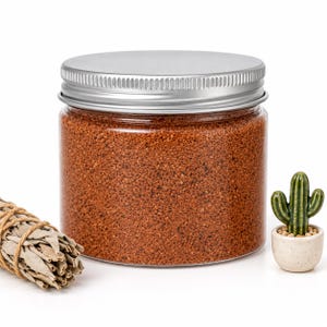 Closed clear jar of Terracotta Clay colored quartz sand in a warm reddish brown, with a silver metal screw lid, next to a bundle of dried white sage tied with twine and a small ceramic cactus in a speckled pot, on a white background.