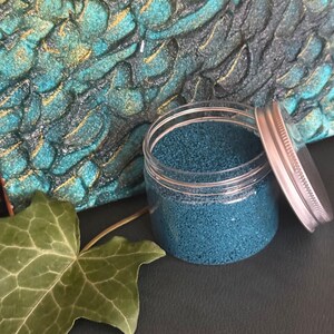 Open jar of Ocean Breeze teal quartz sand with its silver lid leaning against it, placed in front of a textured sand painting in turquoise, dark green and gold, with a green ivy leaf in the foreground
