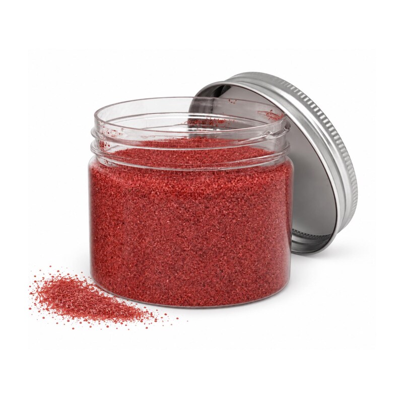 Open clear jar of warm rose red colored quartz sand with its silver metal screw lid leaning behind it and a small pile of the same sand spilled in front, showing the fine grain texture, on a white background.