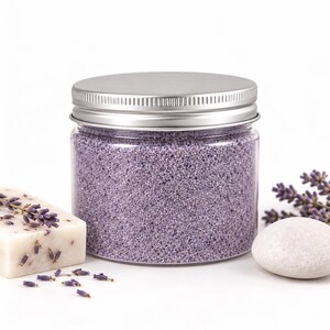 Closed clear jar of Lavender Mist colored quartz sand in a light lavender, with a silver metal screw lid, next to a bar of handmade soap topped with dried lavender, fresh lavender sprigs and a smooth white stone, on a white background.