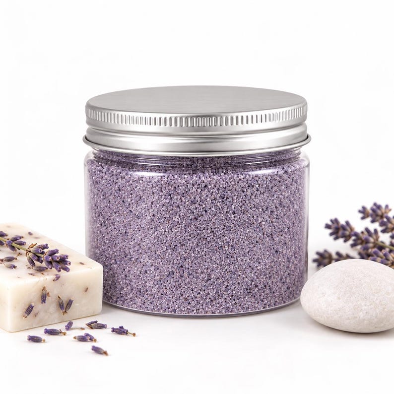 Closed clear jar of Lavender Mist colored quartz sand in a light lavender, with a silver metal screw lid, next to a bar of handmade soap topped with dried lavender, fresh lavender sprigs and a smooth white stone, on a white background.