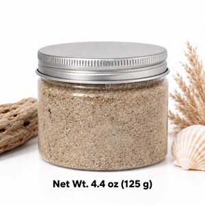 Closed clear jar of Coastal Beige colored quartz sand in a natural sandy beige, with a silver metal screw lid, next to a piece of driftwood, dried pampas grass and a scallop shell, on a white background, with text: Net Wt. 4.4 oz (125 g).