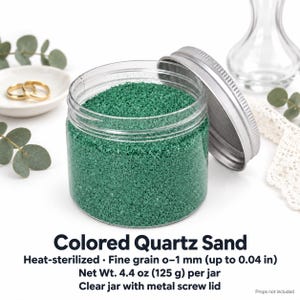 Open clear jar of Lagoon Green colored quartz sand in an emerald green, with the silver metal lid leaning against it, next to two gold wedding rings on a white dish, a clear glass vase, eucalyptus and white lace. Text: Colored Quartz Sand, heat-sterilized, fine grain 0-1 mm (up to 0.04 in), Net Wt. 4.4 oz (125 g) per jar, clear jar with metal screw lid, props not included.