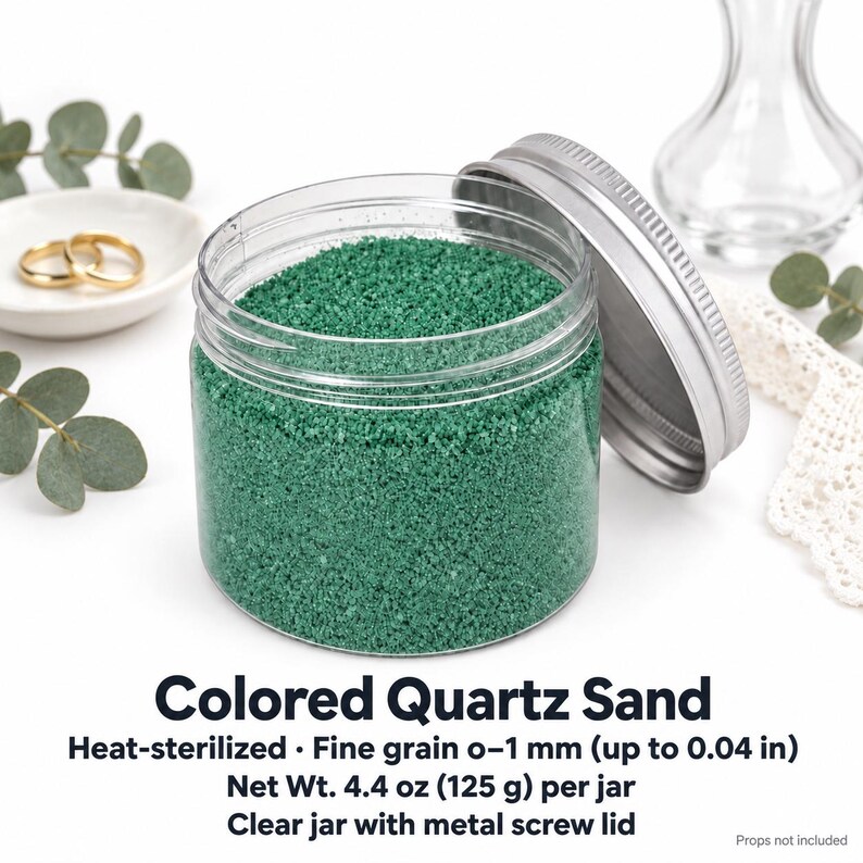 Open clear jar of Lagoon Green colored quartz sand in an emerald green, with the silver metal lid leaning against it, next to two gold wedding rings on a white dish, a clear glass vase, eucalyptus and white lace. Text: Colored Quartz Sand, heat-sterilized, fine grain 0-1 mm (up to 0.04 in), Net Wt. 4.4 oz (125 g) per jar, clear jar with metal screw lid, props not included.