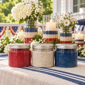 Three closed clear jars of colored quartz sand with silver metal screw lids, red, off-white and royal blue, on a striped linen tablecloth. Behind them are glass jars and candle holders layered with red, white and blue sand, holding white hydrangeas, daisies and lit pillar candles, on a sunny porch with flag bunting.