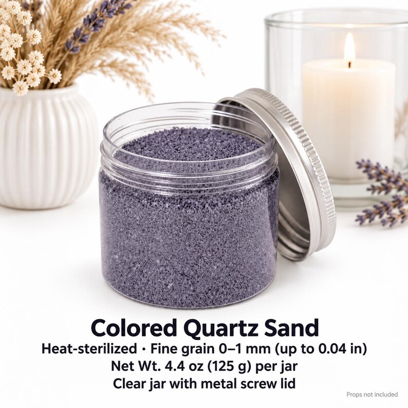 Open clear jar of lavender grey colored quartz sand with the silver metal lid leaning against it, next to a white ribbed vase with pampas grass and dried flowers, a lit white candle in a glass holder and lavender sprigs. Text: Colored Quartz Sand, heat-sterilized, fine grain 0-1 mm (up to 0.04 in), Net Wt. 4.4 oz (125 g) per jar, clear jar with metal screw lid, props not included.