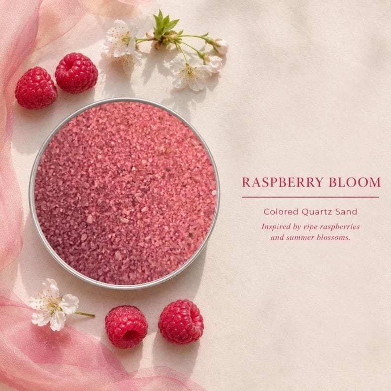 Round tin of Raspberry Bloom colored quartz sand on a cream background, surrounded by fresh raspberries, white blossoms and pink fabric, with text: Raspberry Bloom, Colored Quartz Sand, inspired by ripe raspberries and summer blossoms.
