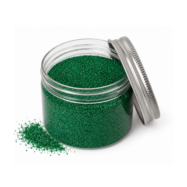 Open clear jar of deep green colored quartz sand with its silver metal screw lid leaning behind it and a small pile of the same sand spilled in front, showing the fine grain texture, on a white background.