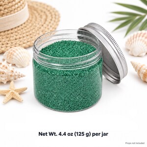 Open clear jar of Lagoon Green colored quartz sand in an emerald green, with the silver metal lid leaning against it, next to a straw hat, seashells, a starfish and a palm leaf. Text:  Net Wt. 4.4 oz (125 g) per jar, props not included.