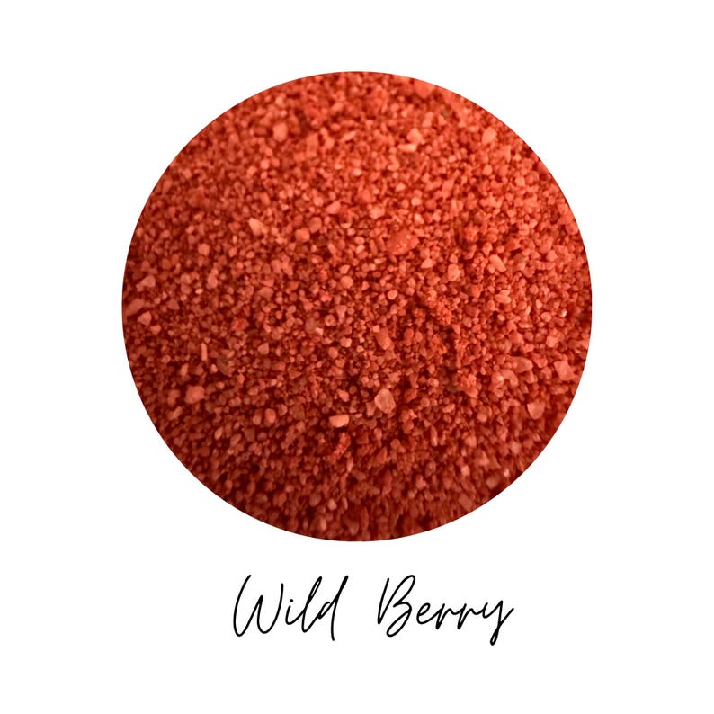 Close-up circle of Wild Berry colored quartz sand showing fine berry red grains, with the color name Wild Berry written in script below, on a white background