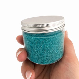 Hand holding a closed clear jar of Ocean Breeze teal blue quartz sand with a silver metal screw lid, showing the jar's small size, on a white background