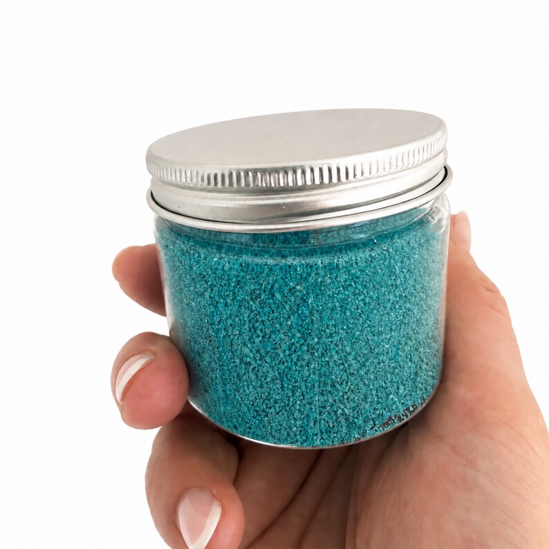 Hand holding a closed clear jar of Ocean Breeze teal blue quartz sand with a silver metal screw lid, showing the jar's small size, on a white background