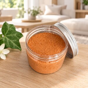 Open jar of orange craft sand with its silver lid leaning on the side, on a wooden table next to an ivy leaf and a white flower