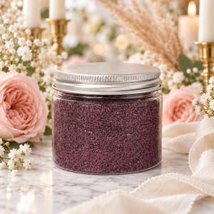 Closed clear jar of Plum Velvet colored quartz sand in a deep plum purple, with a silver metal screw lid on a marble table, surrounded by pink roses, baby's breath, pampas grass, lit white candles in brass holders and cream fabric.