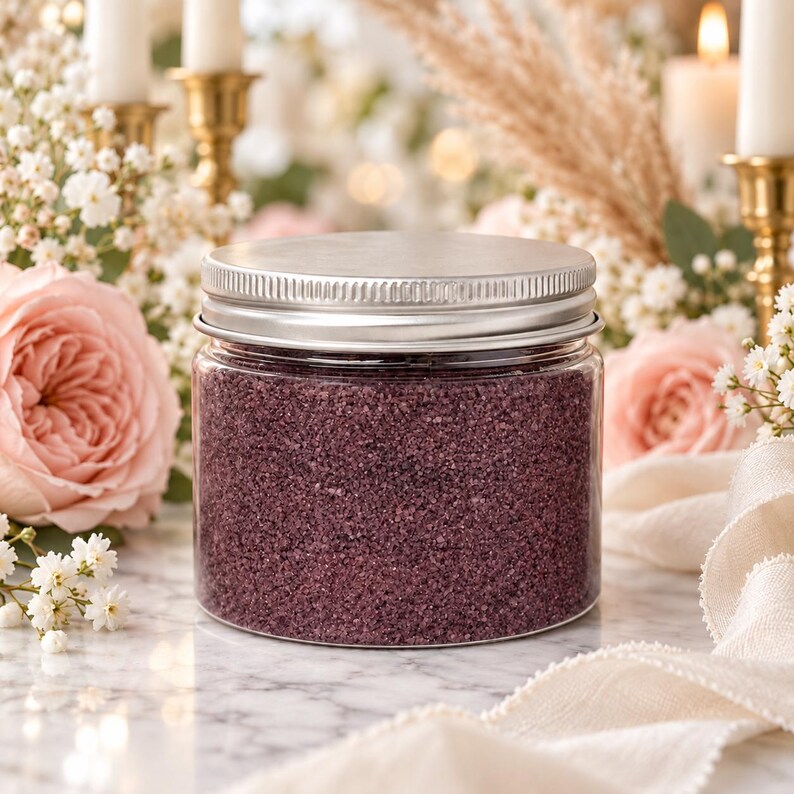 Closed clear jar of Plum Velvet colored quartz sand in a deep plum purple, with a silver metal screw lid on a marble table, surrounded by pink roses, baby's breath, pampas grass, lit white candles in brass holders and cream fabric.
