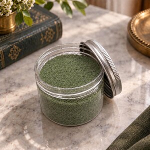 Open jar of Olive Grove olive green quartz sand with its silver lid leaning against it, on a marble surface next to an old green book, a small brass vase with white flowers and a brass tray