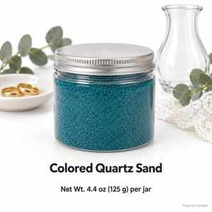 Closed clear jar of deep teal colored quartz sand with a silver metal screw lid, next to two gold wedding rings on a white dish, a clear glass vase, eucalyptus and white lace. Text: Colored Quartz Sand, Net Wt. 4.4 oz (125 g) per jar, props not included.