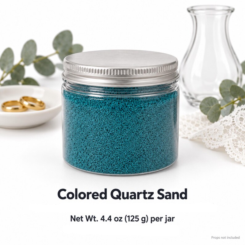 Closed clear jar of deep teal colored quartz sand with a silver metal screw lid, next to two gold wedding rings on a white dish, a clear glass vase, eucalyptus and white lace. Text: Colored Quartz Sand, Net Wt. 4.4 oz (125 g) per jar, props not included.