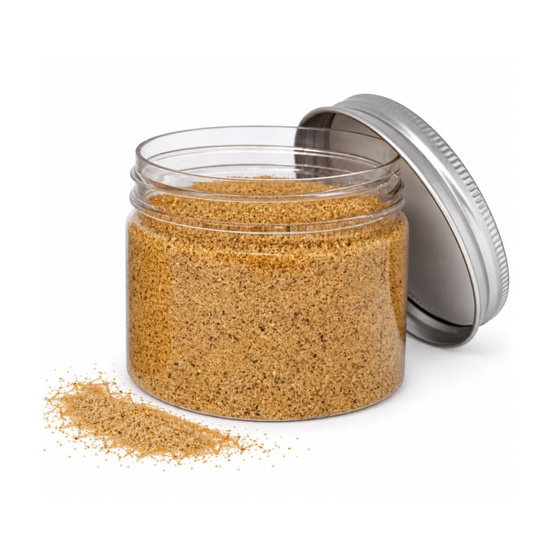 Open clear jar of golden sand-colored quartz sand with its silver metal screw lid leaning behind it, and a small pile of the same sand spilled in front, showing the fine grain texture, on a white background.