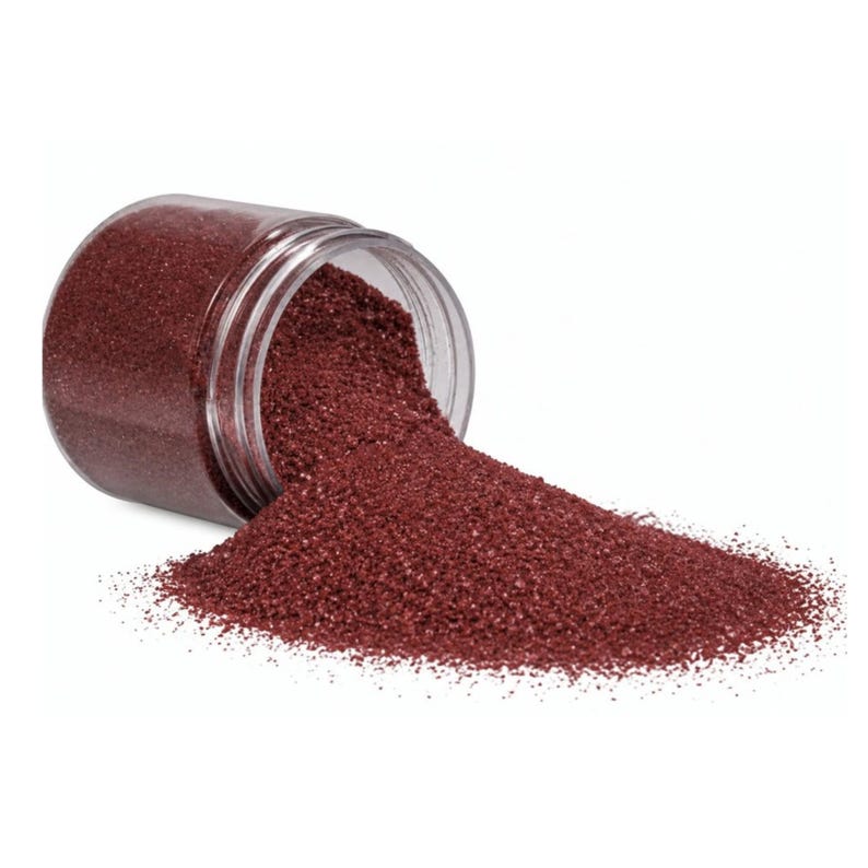 Tipped-over clear jar spilling Wild Berry deep red quartz sand onto a white surface, showing the fine texture of the grains.