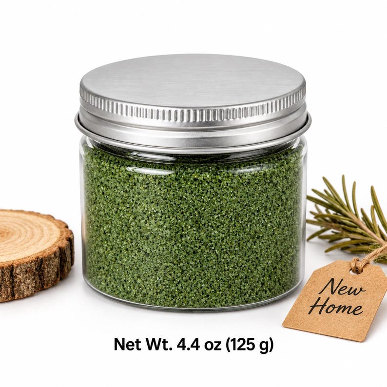 Closed clear jar of moss green colored quartz sand with a silver metal screw lid, next to a wood slice, an evergreen sprig and a kraft paper tag reading New Home, with text: Net Wt. 4.4 oz (125 g).