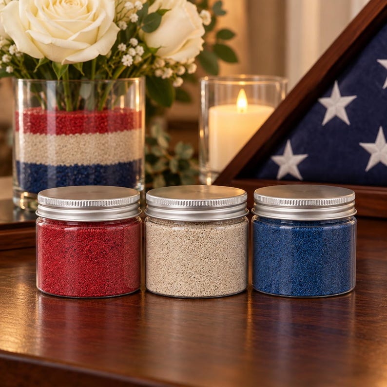 Jars of red, white, and blue craft sand beside a striped sand vase with white roses, a candle, and a folded flag
