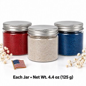 Three closed clear jars of colored quartz sand with silver metal screw lids, in red, off-white and royal blue, next to an American flag pin and sprigs of small white dried flowers, with text: Each Jar, Net Wt. 4.4 oz (125 g).