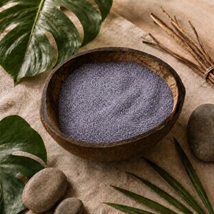 Coconut shell bowl filled with Lavender Mist colored quartz sand in a soft lavender grey, on a linen cloth surrounded by monstera leaves, palm leaves, smooth grey stones and a bundle of dry twigs.