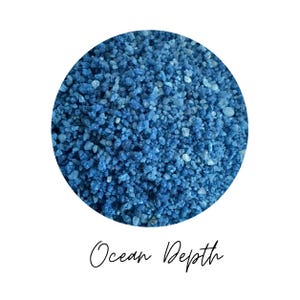 Close-up circle of Ocean Depth colored quartz sand showing blue grains in varied shades, with the color name Ocean Depth written in script below, on a white background