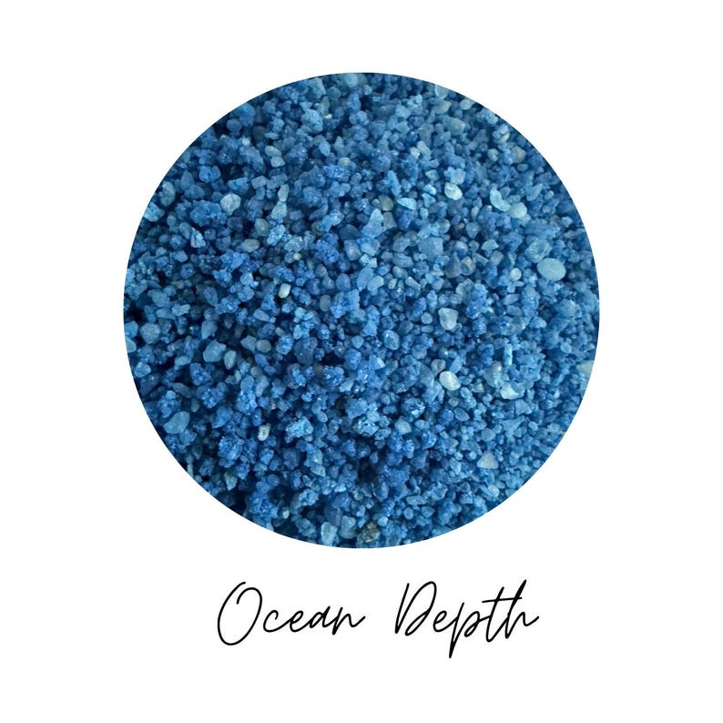 Close-up circle of Ocean Depth colored quartz sand showing blue grains in varied shades, with the color name Ocean Depth written in script below, on a white background