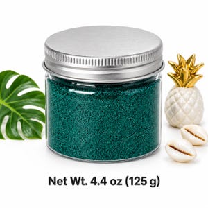 Closed clear jar of deep teal green colored quartz sand with a silver metal screw lid, next to a monstera leaf, a white ceramic pineapple with a gold top and two cowrie shells, with text: Net Wt.