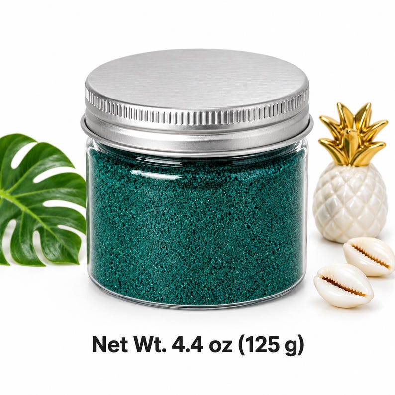 Closed clear jar of deep teal green colored quartz sand with a silver metal screw lid, next to a monstera leaf, a white ceramic pineapple with a gold top and two cowrie shells, with text: Net Wt.