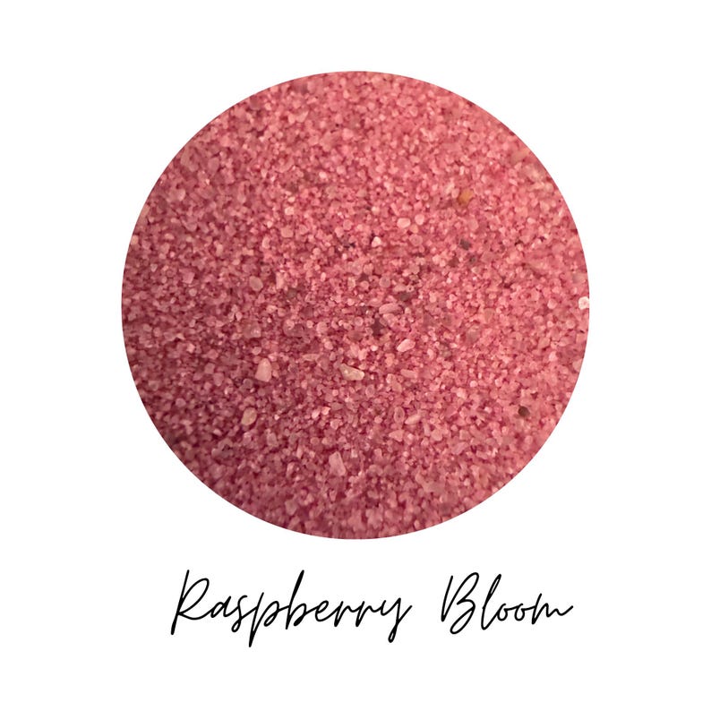 Close-up circle of Raspberry Bloom colored quartz sand showing fine raspberry pink grains, with the color name Raspberry Bloom written in script below, on a white background.