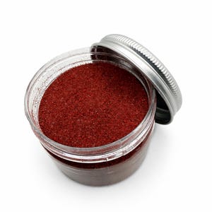Top view of an open jar of Wild Berry red quartz sand with the silver metal lid leaning against its side, on a white background.
