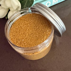 Open jar of Amber Gold craft sand with silver lid on a dark table next to white flowers