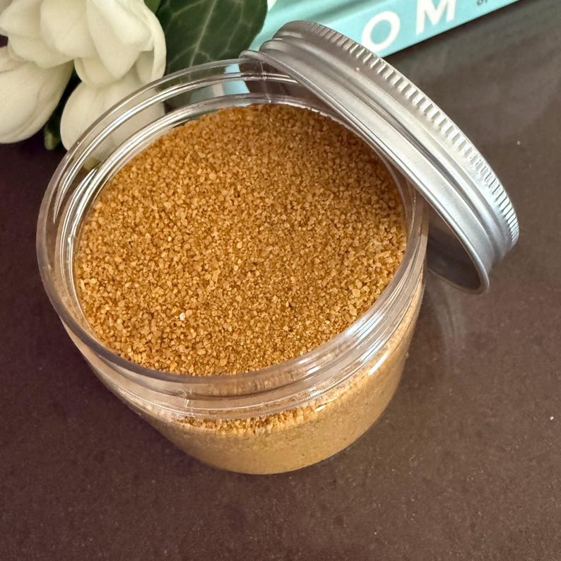 Open jar of Amber Gold craft sand with silver lid on a dark table next to white flowers